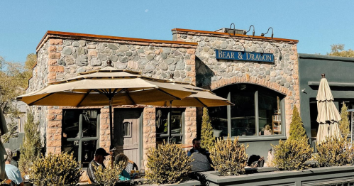 A stone building with a sign reading "Bear & Dragon," featuring outdoor seating under umbrellas and greenery.
