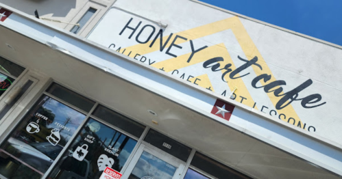 Exterior of Honey Art Cafe, featuring a sign with "Gallery + Cafe + Art Lessons" and large windows displaying various icons.
