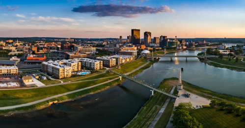 Aerial view of a city skyline with buildings, a river, and a bridge at sunset. Green spaces line the water's edge.