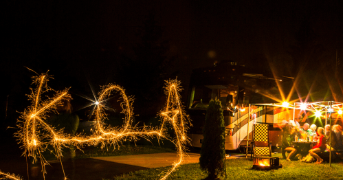 A nighttime scene with a camper and people celebrating, writing "KOA" with sparklers.