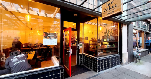A cozy bakery entrance with large windows, warm lighting, and a welcoming red door. People are visible outside.