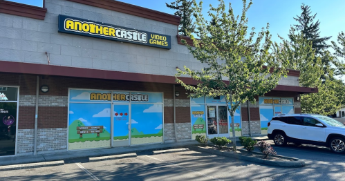 Exterior of a video game store named "Another Castle," featuring colorful window graphics and a parked white SUV.