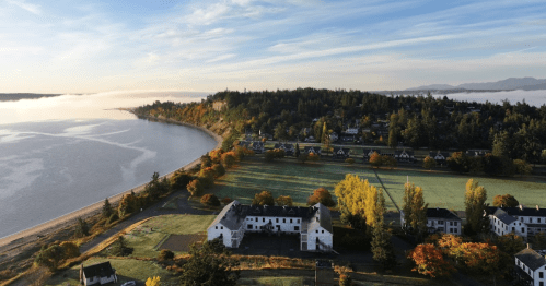 Aerial view of a coastal landscape with fog, trees in autumn colors, and a historic building near the shore.