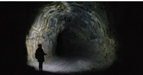A person stands in a dark cave, illuminated by a flashlight, revealing textured rock walls and a distant opening.
