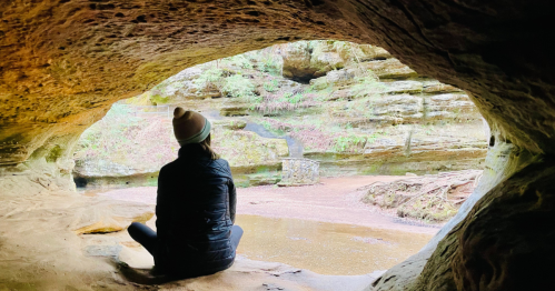 A person sits in a cave, looking out at a serene landscape with water and greenery.