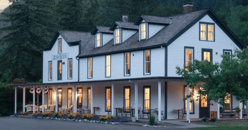A charming white house with multiple windows, surrounded by trees and flowers, illuminated in the evening light.