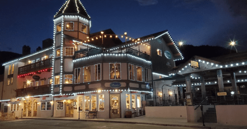 A charming, illuminated building with festive lights at night, featuring a tower and outdoor seating.
