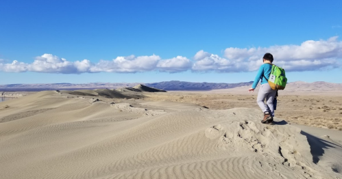 A person walks along a sandy ridge in a desert landscape under a blue sky with fluffy clouds.