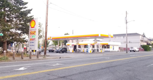 A Shell gas station on a street, with fuel pumps and a sign displaying prices, surrounded by trees and buildings.