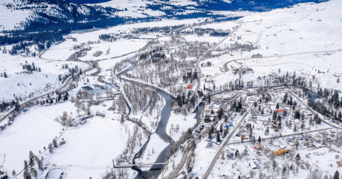 Aerial view of a snowy landscape with a winding river and small town surrounded by trees and mountains.