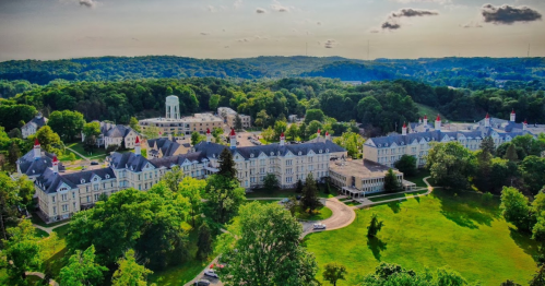 Aerial view of a large, historic campus surrounded by lush greenery and rolling hills under a partly cloudy sky.