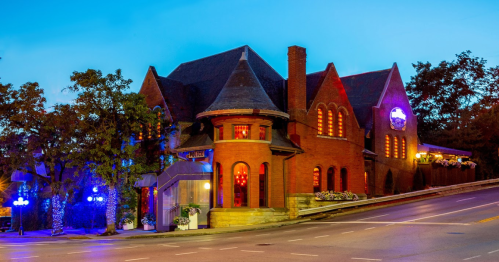 A charming brick building with a steep roof, illuminated at dusk, set on a quiet street with trees nearby.