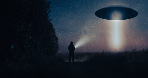 A person stands in a dark field, holding a flashlight, as a UFO hovers above, emitting a beam of light.