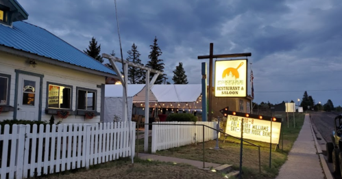 A restaurant and saloon with a sign, outdoor seating, and a cloudy sky in the background. White picket fence visible.