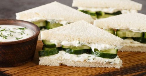 Cucumber sandwiches with creamy filling, served on a wooden board alongside a bowl of dip.