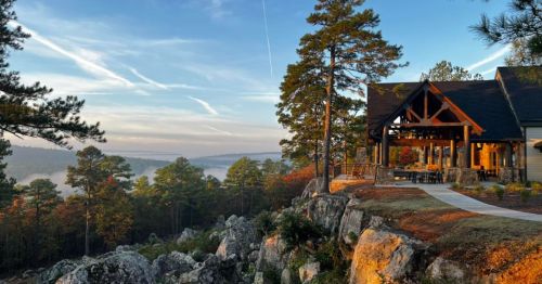 A scenic view of a house on a cliff, surrounded by trees and misty mountains under a clear blue sky.