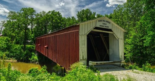 A red covered bridge, Code Mill Bridge, built in 1864, surrounded by lush greenery and a calm river.