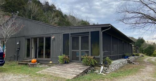 A modern, dark wooden house with a screened porch, surrounded by greenery and a gravel path.