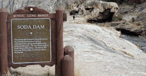 A scenic marker for Soda Dam, with a rocky landscape and a person walking in the background near a water feature.