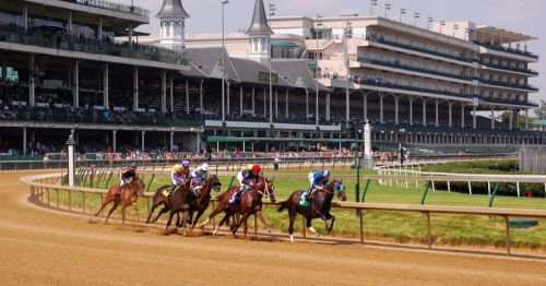 Horses racing on a dirt track with a grandstand in the background, filled with spectators on a sunny day.