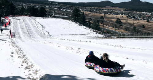 Two people slide down a snowy hill on colorful inner tubes, with a scenic landscape in the background.