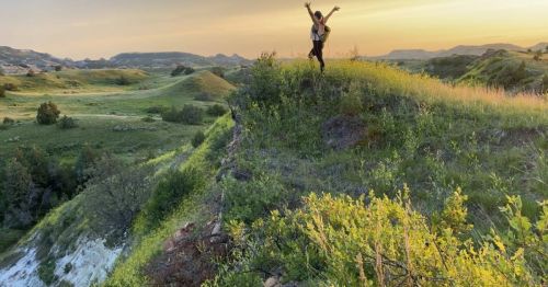 A person stands on a grassy hilltop at sunset, arms raised, overlooking a scenic landscape of rolling hills.