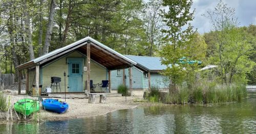 A cozy cabin by the water, surrounded by trees, with kayaks in front and a peaceful outdoor setting.