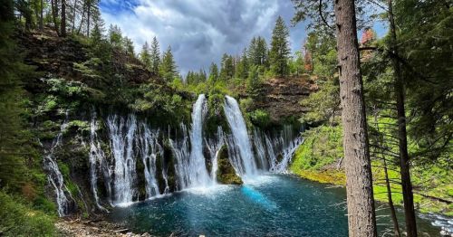A scenic view of a waterfall cascading into a clear blue pool, surrounded by lush green trees and a cloudy sky.