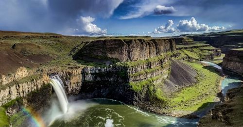 A stunning waterfall cascades into a river, surrounded by lush green cliffs and a vibrant blue sky with clouds.