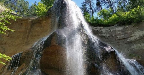 A cascading waterfall flows down a rocky cliff surrounded by lush green trees under a clear blue sky.