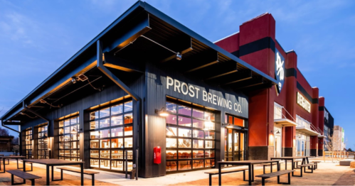 Exterior of Prost Brewing Co. featuring large windows, modern architecture, and outdoor seating at dusk.