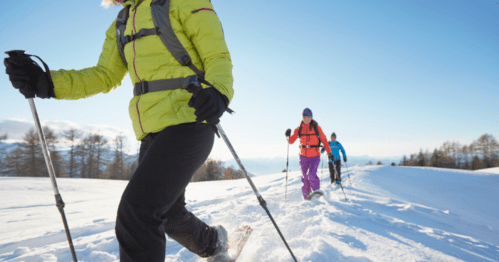 Three people hiking in the snow, wearing colorful winter gear and using trekking poles against a clear blue sky.