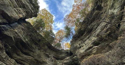 View looking up from a rocky canyon, framed by tall cliffs and trees with autumn foliage against a blue sky.