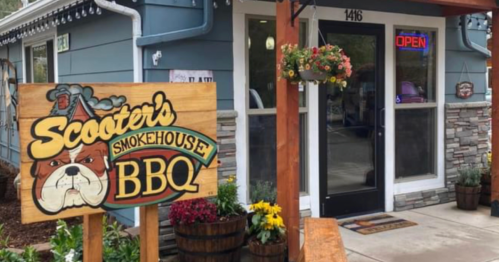 Sign for Scooter's Smokehouse BBQ outside a blue building with an "Open" sign and flower planters.