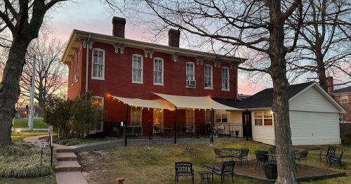 A red brick building with a white extension, surrounded by trees and outdoor seating, under a colorful sunset sky.