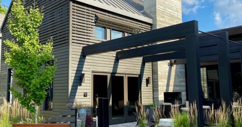 Modern house with wooden slats, large windows, and a black pergola, surrounded by greenery and decorative plants.