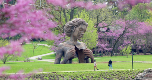 A large wooden statue of a woman stands in a park, surrounded by blooming trees and people walking along a path.