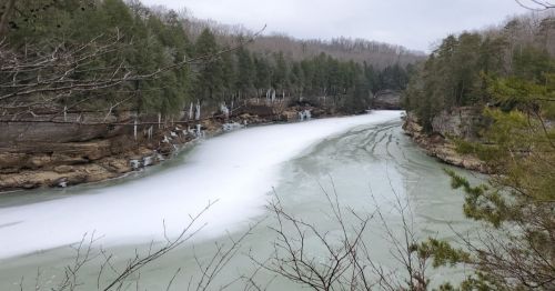 A serene river scene with partially frozen water, surrounded by trees and rocky cliffs under a cloudy sky.