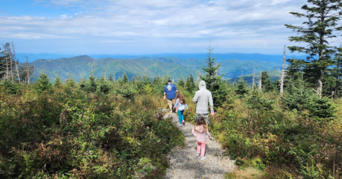 A family walks along a trail in the mountains, surrounded by greenery and distant blue hills under a cloudy sky.