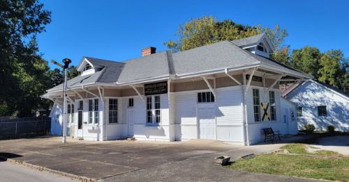 Historic white building with a gray roof, featuring large windows and a sign, set against a clear blue sky.