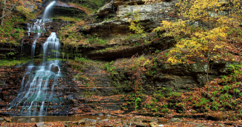 A serene waterfall cascading down rocky cliffs, surrounded by autumn foliage and fallen leaves.