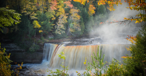 A scenic waterfall surrounded by vibrant autumn foliage and mist rising from the water.