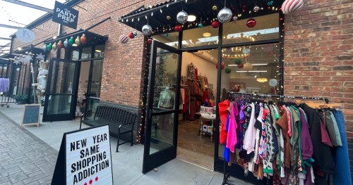 A festive storefront with holiday decorations, featuring a clothing rack outside and a sign promoting shopping.