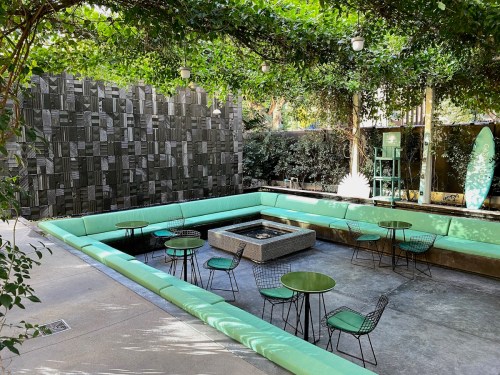 A modern outdoor lounge area with green seating, tables, and a fire pit, surrounded by lush greenery.