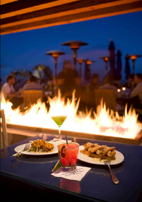 A cozy outdoor dining scene with two plates of food, colorful cocktails, and a warm fire in the background at dusk.