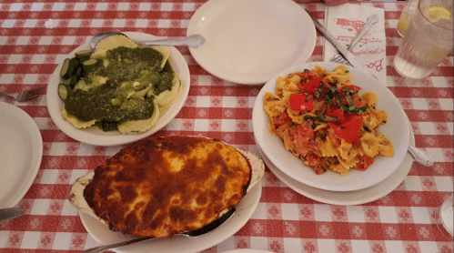Three plates of Italian food: pesto ravioli, pasta with tomatoes, and a baked cheese dish on a checkered tablecloth.