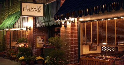 A cozy restaurant entrance with a sign reading "Food & Friends," featuring outdoor seating and warm lighting.