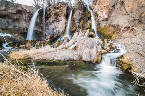 A serene landscape featuring cascading waterfalls over rocky terrain, surrounded by dry grass and trees.