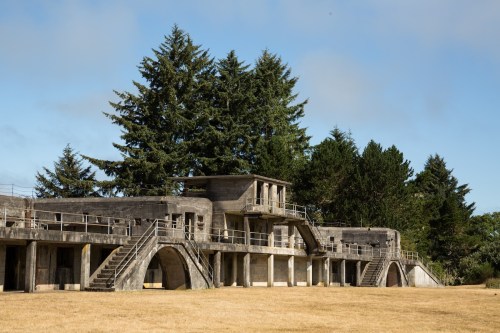 Abandoned concrete structure with multiple levels, surrounded by tall trees and a grassy field under a clear sky.