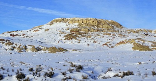 A snow-covered landscape with a prominent, layered rock formation under a clear blue sky.